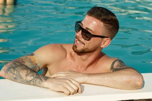 Close up portrait of a handsome bearded man in sunglasses smiling looking away while relaxing at the swimming pool. Happy man resting after swimming. Summer, trip, recreation, leisure concept