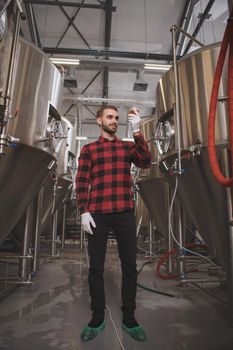 Low angle vertical shot of a handsome bearded man tasting beer at microbrewery, standing between metal beer tanks