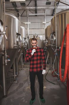 Vertical full length shot of a professional brewer standing near beer tanks at his microbrewery, sipping fresh beer