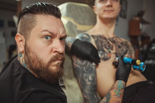 Bearded professional tattoo artist making angry face to the camera, while working with his male client, tattooing him on chest. Tattooist making funny face while working