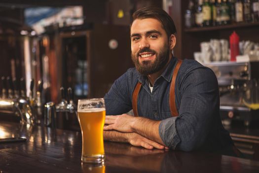 Handsome bearded bartender smiling joyfully to the camera, leaning on the bar counter, serving delicious beer at his pub, copy space. Brewer offering tasty craft beer at his restaurant
