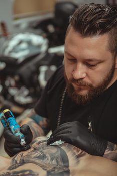 Vertical shot of a bearded professional tatoo artist concentrating, while working at his studio. Cropped shot of a tattooist drawing tattoo on chest of a client