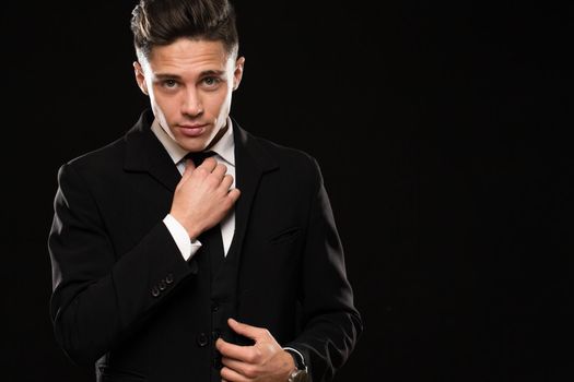Young handsome man wearing elegant black suit adjusting his tie looking to the camera seductively on black background copyspace lifestyle macho boyfriend sexuality tempting confidence power