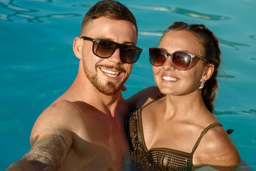 Lovely young couple relaxing in outdoor pool during summer vacation. Happy boyfriend and girlfriend taking selfie while resting in the swimming pool. Gorgeous woman and her handsome man smiling joyfully