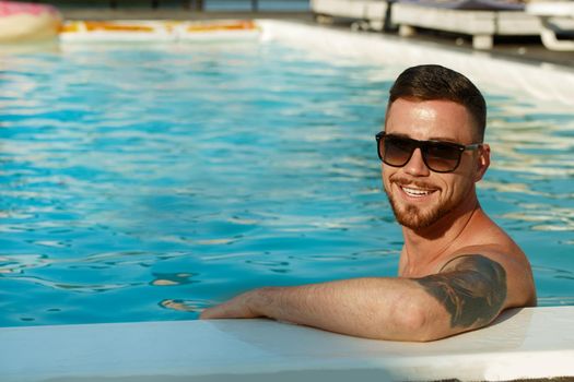 Cheerful handsome bearded man with tattoos smiling to the camera, while relaxing in the swimming pool, copy space. Happy young man enjoying swimming in the pool on a hot sunny day