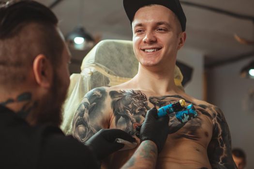 Handsome cheerful young man smiling, talking to his tattoo artist while getting inked at salon. Attractive male client getting chest tattoo by professional tattooist. Service, consumerism concept