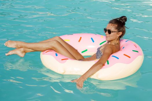 Young happy woman enjoying day at the pool swimming on inflatable donut, copy space. Beautiful woman tanning while swimming on donut float at the resort pool. Recreation, enjoyment, summer concept.