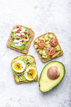 Avocado toasts with egg, tomatoes, seasonings and a half of whole avocado over white stone background. Healthy breakfast avocado sandwiches with different toppings, top view, close-up.