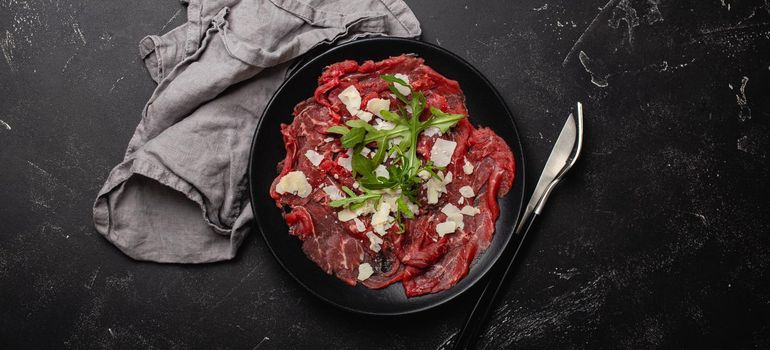 Italian cold meat appetizer Beef carpaccio with parmesan cheese and arugula on black plate with fork and knife on dark stone concrete rustic background flat lay from above .