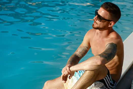 Handsome tattooed bearded man relaxing by the pool on a hot sunny day. Attractive young man enjoying hos summer vacation, resting at the poolside, copy space