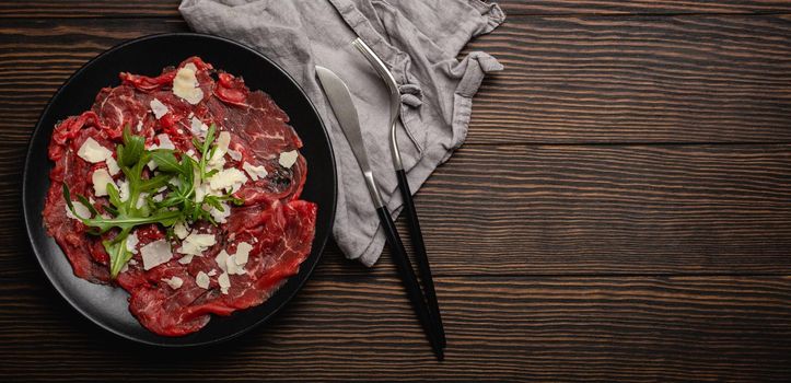 Italian cold meat appetizer Beef carpaccio with parmesan cheese and arugula on black plate with fork and knife on dark brown wooden rustic background flat lay from above space for text.