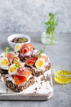 Close-up of sandwich with dark rye bread, cream cheese, salmon, onion, capers, boiled egg on white wooden cutting board, concrete background. Traditional Scandinavian open sandwich Smorrebrod.