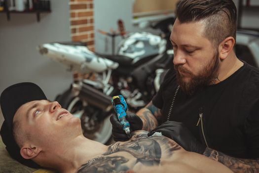 Cropped shot of a handsome young man getting tattooed on chest by professional tattoo artist. Bearded male tattooist working at his studio, making tatoo on chest of his client