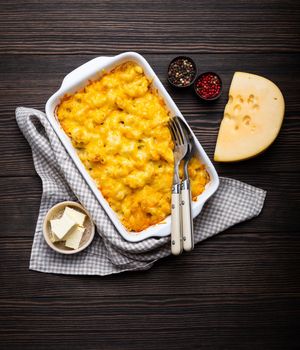 Close-up of baked mac and cheese in white casserole on rustic wooden background, with seasonings, butter, spoon and fork, top view. Pasta with cheesy sauce, American/English comfort food