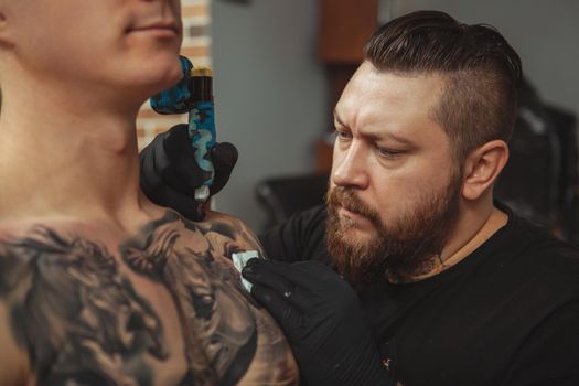 Close up of a concentrated bearded tattoo artist working, tattooing shoulder of a male client. Professional tattooist looking focused, copy space
