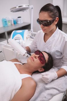 Vertical shot of a professional beautician and female client wearing protective glasses during facial hair laser removal procedure