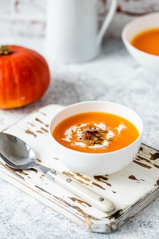 Homemade pumpkin cream soup served in white ceramic bowl on white table with spoon decorated with whole pumpkin, angle view, selective focus. Autumn cozy comfort food .