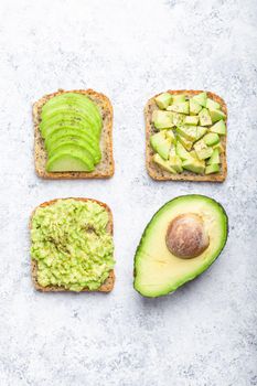 Different types of avocado toasts and a half of whole avocado over white stone background, concept. Ways of making and serving delicious healthy breakfast avocado sandwich, top view, close-up.