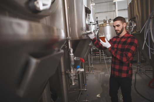 Male brewer tasting freshly brewed beer at his brewery, metal tanks on background, copy space