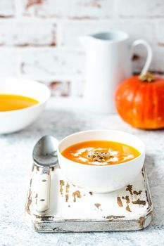 Homemade pumpkin cream soup served in white ceramic bowl on white table with spoon decorated with whole pumpkin, angle view, selective focus. Autumn cozy comfort food .