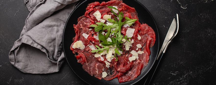 Italian cold meat appetizer Beef carpaccio with parmesan cheese and arugula on black plate with fork and knife on dark stone concrete rustic background flat lay from above .
