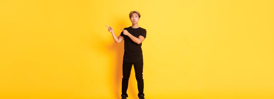 Full-length of sad and miserable asian guy in black clothes, pouting, pointing fingers upper right corner, standing yellow background.