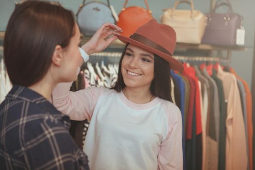 Beautiful cheerful young woman trying on a hat at clothing store, talking to her friend, copy space. Two young women enjoying shopping for clothing and accessories together