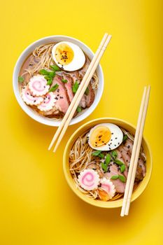 Two bowls of Japanese noodle soup ramen with meat broth, sliced pork, narutomaki, egg with yolk on pastel yellow background. Traditional dish of Japan, top view, close-up, concept