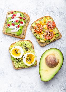 Avocado toasts with egg, tomatoes, seasonings and a half of whole avocado over white stone background. Healthy breakfast avocado sandwiches with different toppings, top view, close-up.