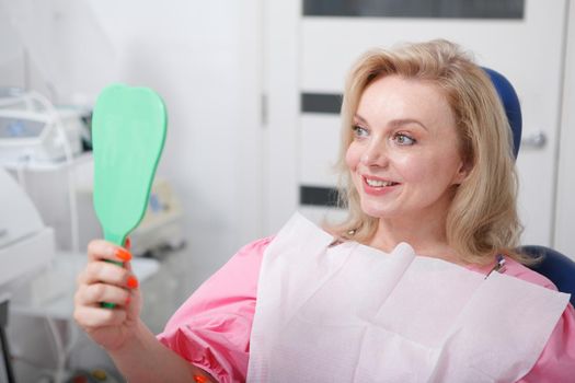 Cheerful mature woman smiling to the mirror she is holding, checking teeth after dental treatment