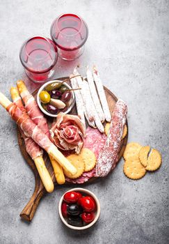 Variety of cold meat cuts and appetizers, red wine, prosciutto, jamon, salami slices, sausage, grissini, olives. Assorted mix of meat on rustic wooden board, grey stone background, top view, close-up.