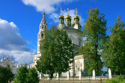 A large old white stone church against the backdrop of large trees and a blue sky.