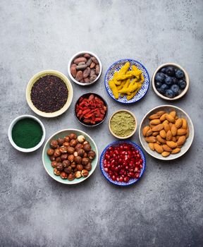 Set of different superfoods in bowls on stone gray background: spirulina, goji berry, cocoa, matcha green tea powder, quinoa, chia seeds, blueberries, nuts for happy healthy living, top view, close up