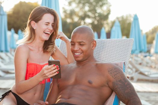 Beautiful happy multiethnic couple laughing, using smart phone together at the beach, copy space. Love, travel, vacation concept