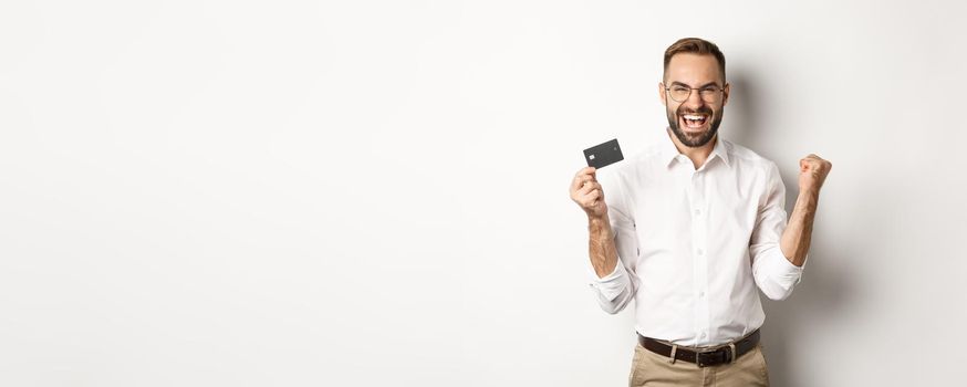 Satisfied handsome man showing credit card and rejoicing, winning something or triumphing, standing over white background.