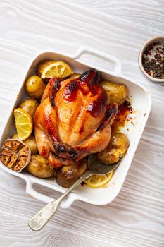 Close-up of baked or roasted whole juicy crispy organic chicken with potatoes, garlic, lemon in white casserole dish with serving spoon on white wooden background, top view