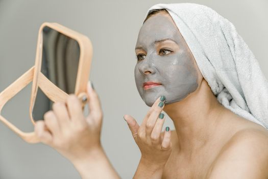 Girl puts gray cosmetic clay mask on her face while looking in mirror, her hair and body are wrapped in towel. Close-up