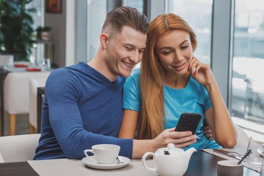 Handsome man smiling, embracing his gorgeous girlfriend, showing her something on smart phone. Beautiful couple having breakfast at the cafe surfing internet. Social media, communication concept