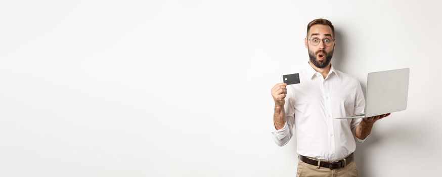 Online shopping. Surprised man holding laptop and credit card, shop internet store, standing over white background.