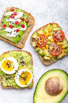 Avocado toasts with egg, tomatoes, seasonings and a half of whole avocado over white stone background. Healthy breakfast avocado sandwiches with different toppings, top view, close-up.