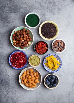 Set of different superfoods in bowls on stone gray background: spirulina, goji berry, cocoa, matcha green tea powder, quinoa, chia seeds, blueberries, nuts for happy healthy living, top view, close up