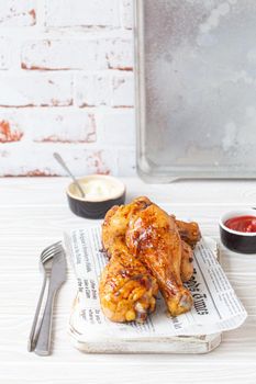 Roasted chicken legs drumsticks on wooden board with newspaper sheet served with ketchup and mayonnaise on rustic white wooden table, angle view