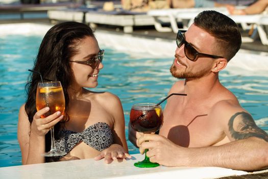 Young loving couple enjoying cocktails together at the swimming pool. Cheerful young woman drinking with her boyfriend while on summer vacation. Recreation, tourism, travelling concept