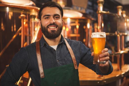 Happy bearded male brewer smiling to the camera joyfully, holding glass of delicious freshly brewed beer. Cheerful beermaker enjoying working at his brewery. Small business owner concept