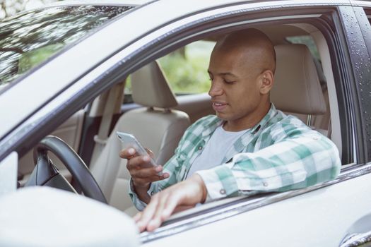 Young African handsome man using his smart phone, sitting in a car on countryside road, copy space. Attractive male driver using online map, travelling by car