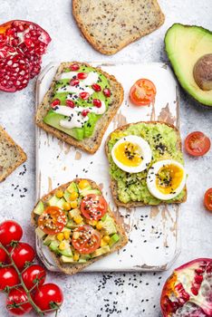 Avocado toasts with egg, tomatoes, seasonings on white wooden rustic cutting board, stone background. Ingredients for healthy breakfast avocado sandwiches with different toppings, top view, close-up.