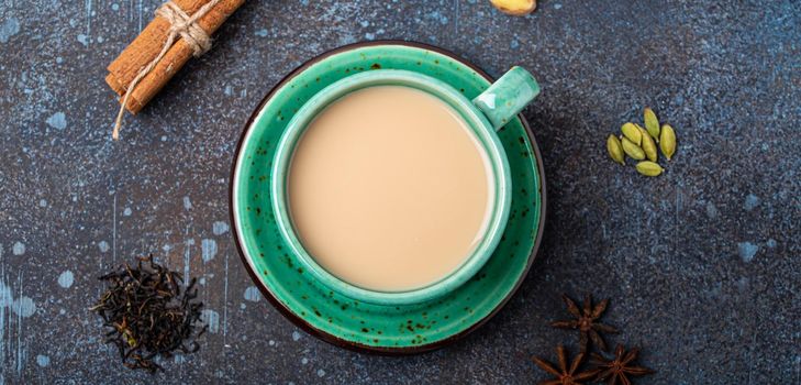 Healthy Indian beverage masala chai - tea hot drink with milk and spices in rustic green teacup with ingredients for making masala chai from above on concrete blue background