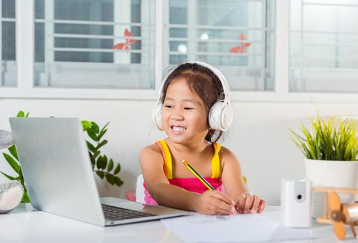 Asian child girl studying video conference distant education at home. Little kid preschool wear headphones sit at desk use laptop computer and communicates on Internet online video call with teacher