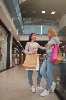 Vertical full length shot of two female friends carrying shopping bags, talking at the mall