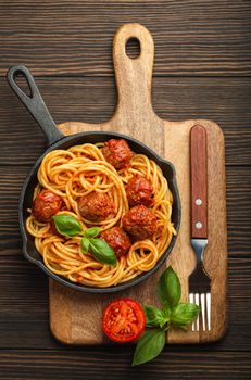 Top view of delicious pasta with meatballs, tomato sauce and fresh basil in cast iron rustic vintage pan served on cutting board, wooden background. Tasty homemade meatballs spaghetti